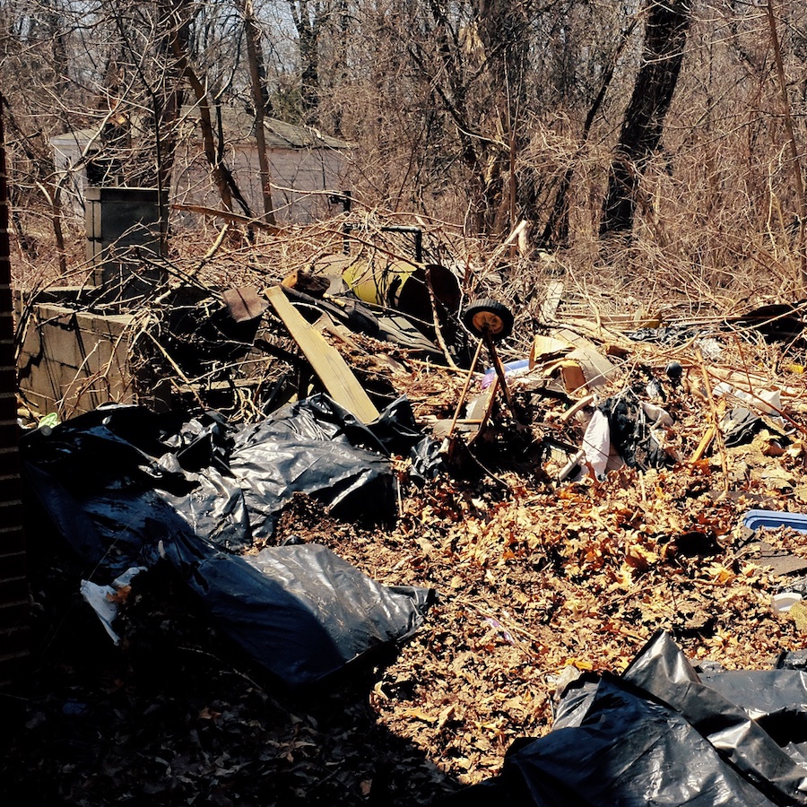 Photograph of driveway area behind house. A concrete grill area is visible. There is huge amount of brush and construction refuse in a pile.