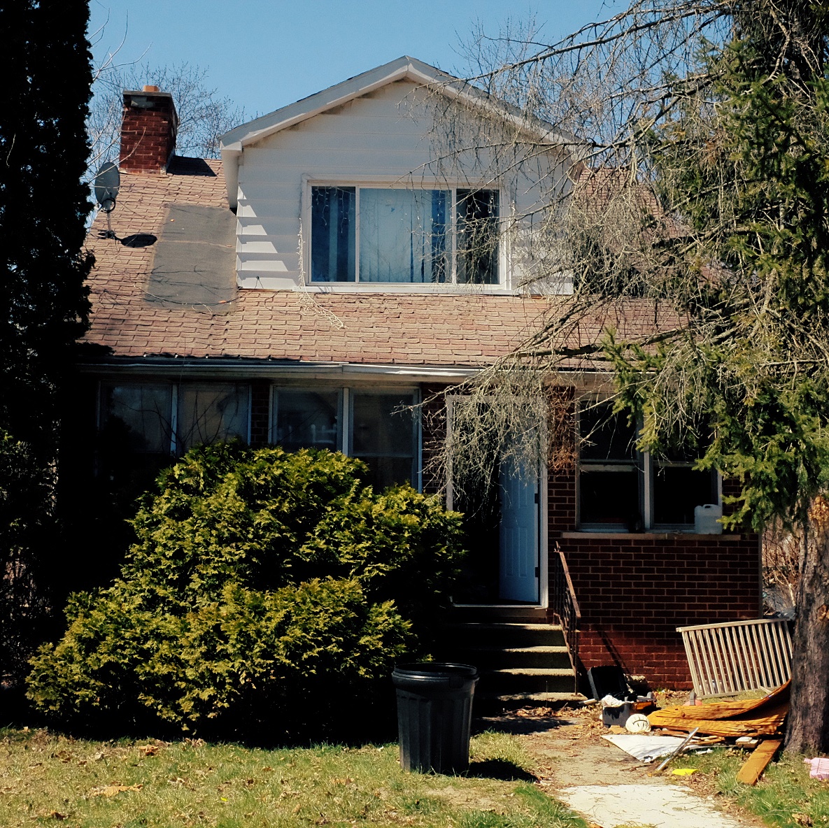 Photograph of a brick bungalo from the front. There's a bush on the left. The front door is open. There is a trash pile on the right at the base of a tree.