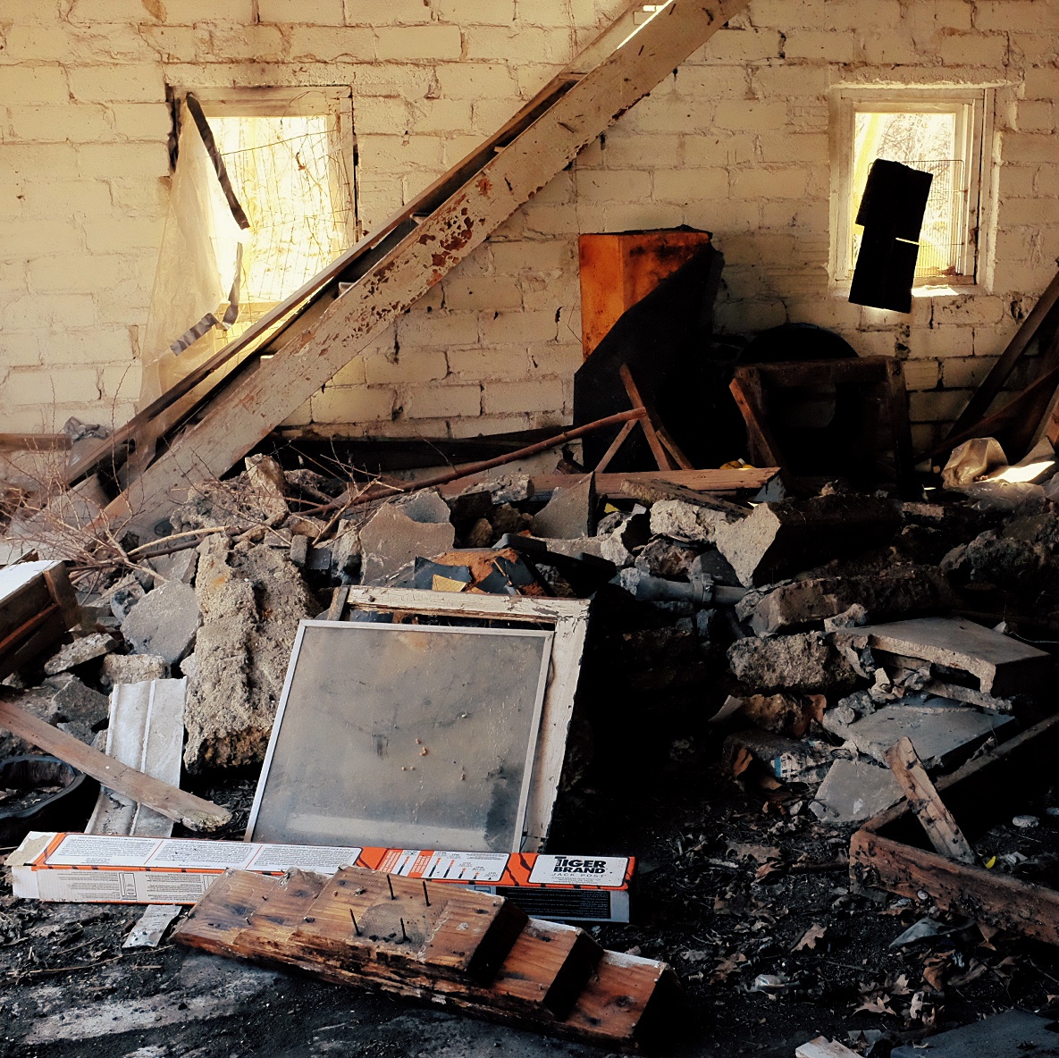 Photograph from inside a cinderblock two car garage. There is a large pile of broken concrete. Stairs in the background. Two open windows in the wall.