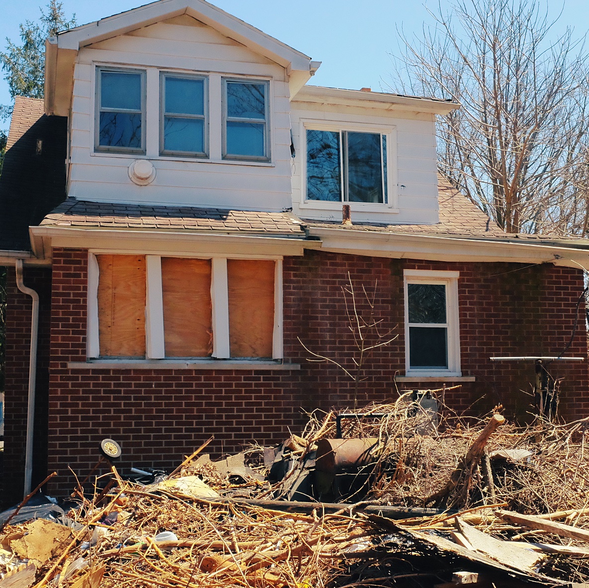 Photograph of the rear of the house showing multiple intact windows on the second floor, both boarded up and intact windows on the main floor. There is huge amount of brush and construction refuse in a pile.