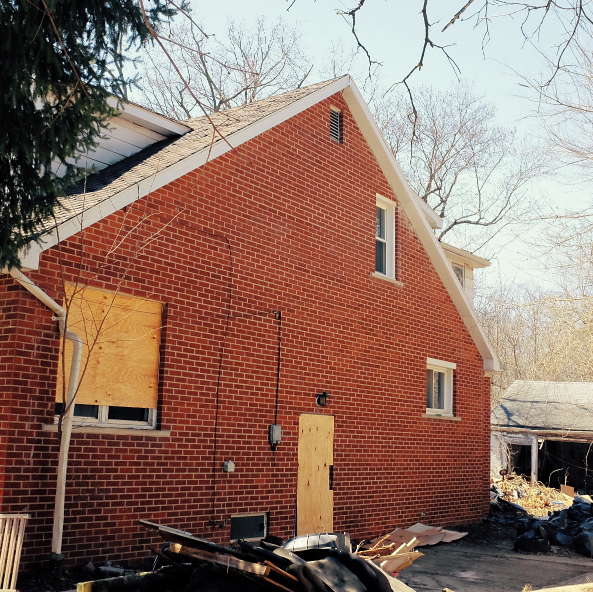 Photograph of right side of the house showing two open windows, one boarded up window, and a boarded up door. There is some garbage in the driveway in piles.