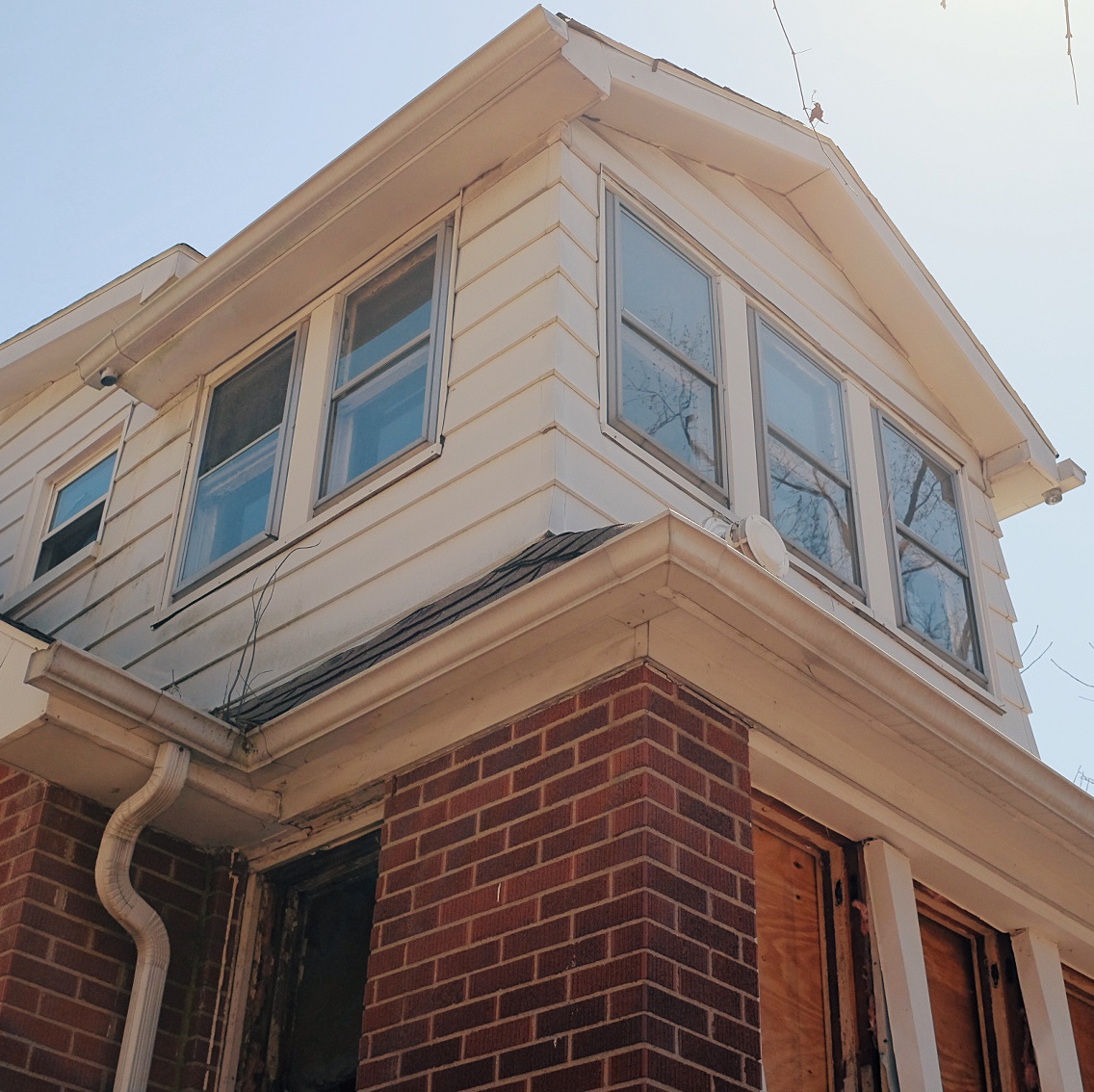 Photograph of the rear, left corner of the house with an upward angle showing a small room on the second floor. Boarded up windows can be seen on the main floor.